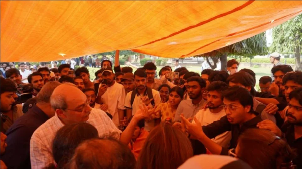 a dense crowd gathered beneath a bright orange canvas tarp at Jamia Millia Islamia (JMI) university, showing a jmi administrative official engaging in negotiation discussion with protesting students