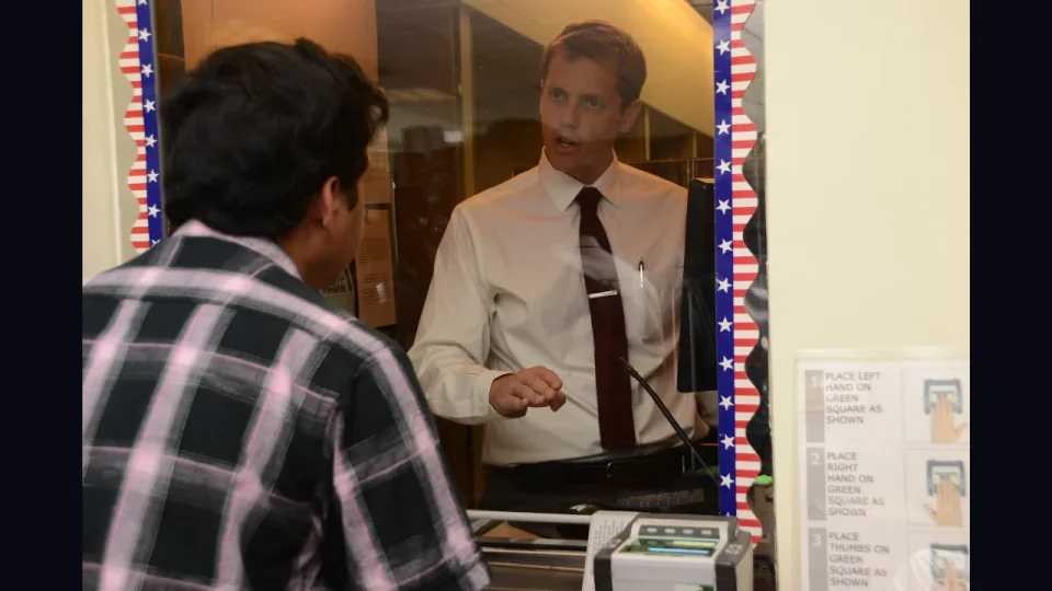  A US visa officer talking to an applicant at an official processing window equipped with a biometric fingerprint scanner and instructional signs.